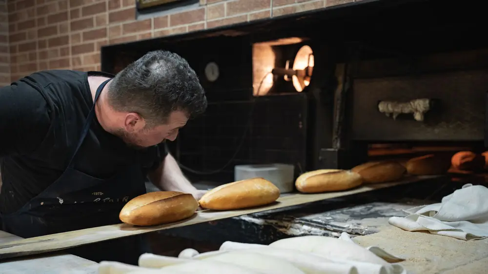 A baker in a black apron sliding fresh loaves into a brick oven on bake day